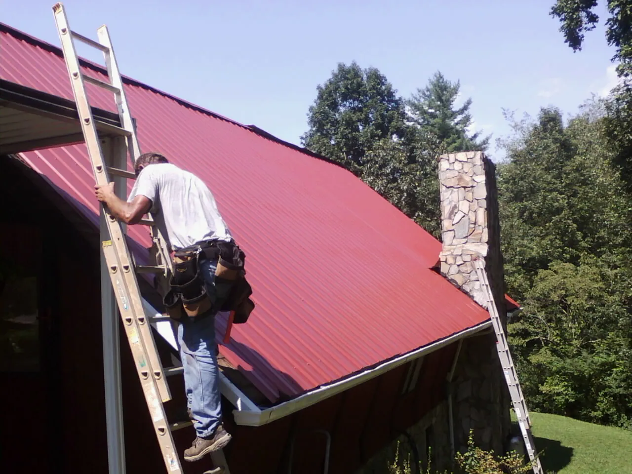 Roofer on a ladder beside a red metal roof and stone chimney — Soffit & Fascia Repair by Roofing Brothers in Clintondale